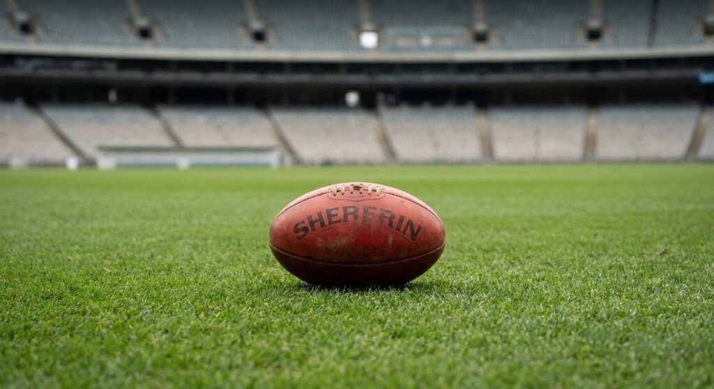 A red Australian rules football sits on green grass. An empty stadium with tiered seating is blurred in the background